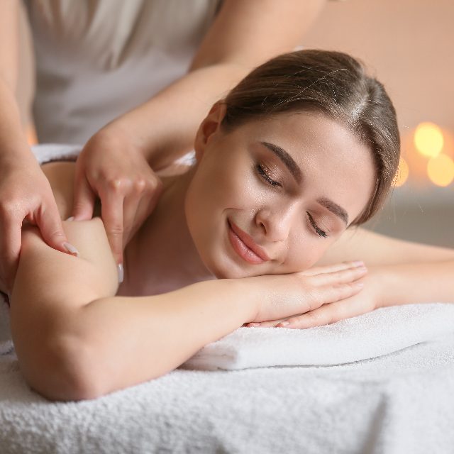 A therapist massages the shoulders of a smiling client lying face down on a towel-covered table
