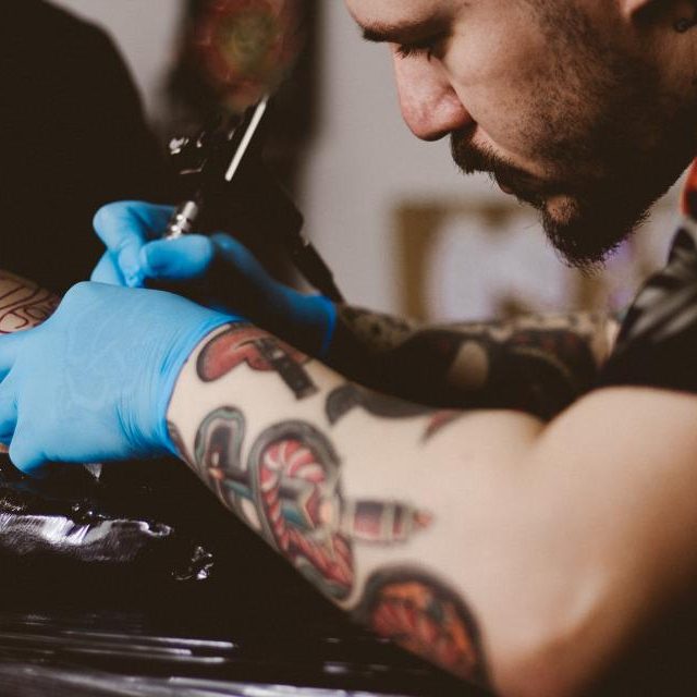 A tattoo artist in blue gloves works on a client's forearm, which already carries colorful traditional tattoos